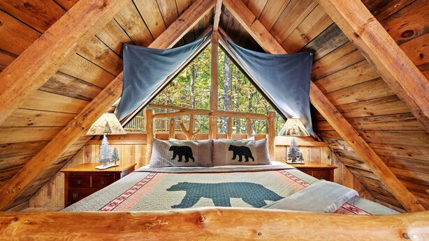 Un dormitorio acogedor en el ático con techo de madera, una cama con edredón de temática de oso y vistas a árboles a través de una ventana.