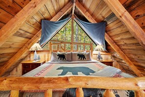 A cozy attic bedroom with a wooden ceiling, a bed with a bear-themed bedspread, and a view of trees through a window.