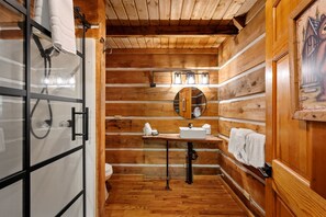 A wooden cabin bathroom with a glass shower enclosure, a sink with a mirror, and a towel rack. - Rustic Cabin Retreat in Sky Harbor | Sleeps 8-10 | Family-Friendly Fun! (Sevierville)