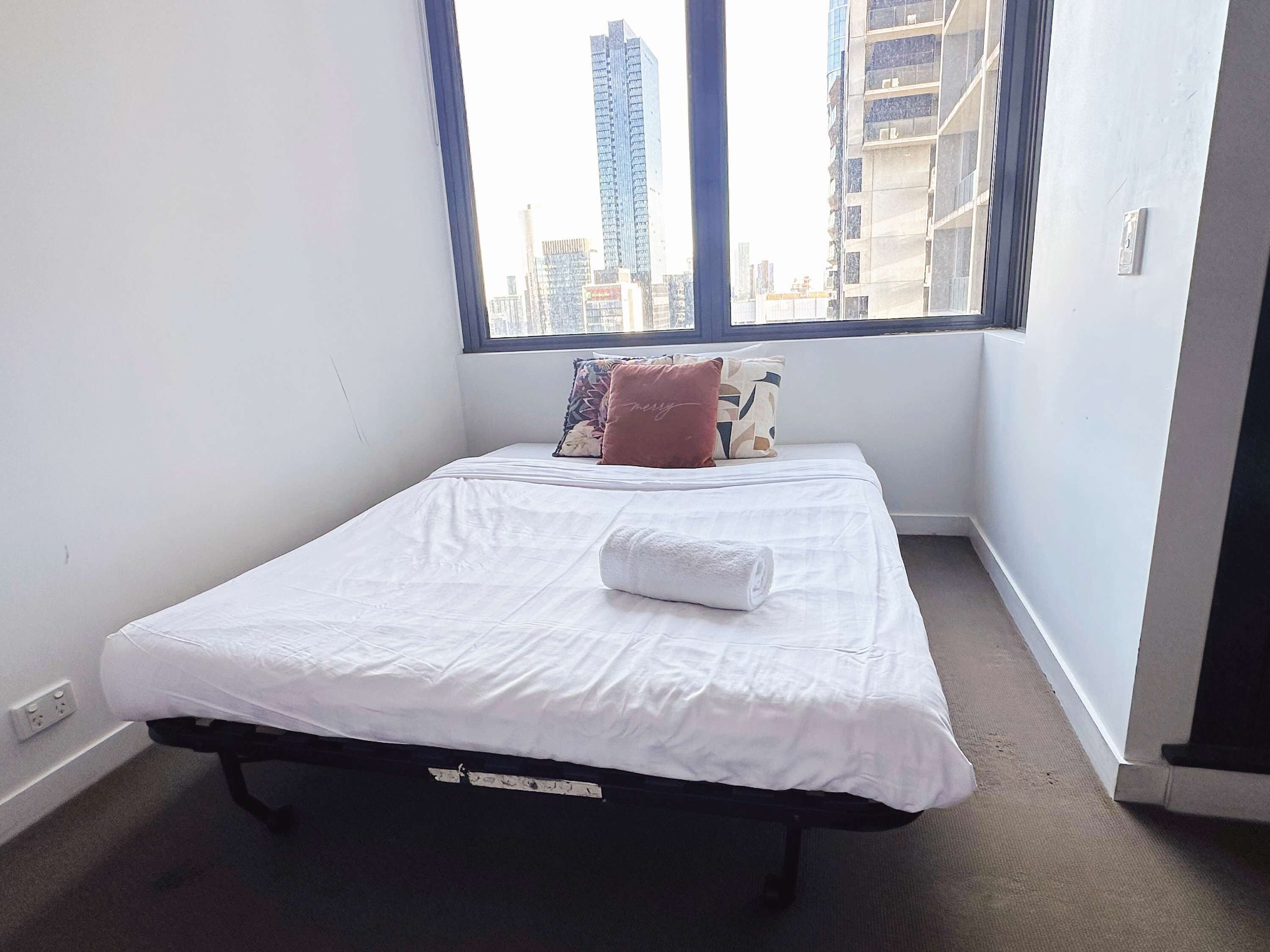 Una cama individual con ropa blanca y una toalla enrollada, una ventana con vista a edificios altos y una pared blanca lisa.