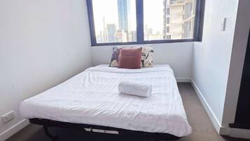 Una cama individual con ropa blanca y una toalla enrollada, una ventana con vista a edificios altos y una pared blanca lisa.