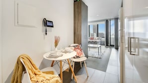 A modern dining area with a table set for two, a chair with a yellow blanket, a mounted phone, and a view of a balcony with a cityscape. - Cozy Studio Apt Near Southern Cross & City Views (Melbourne)