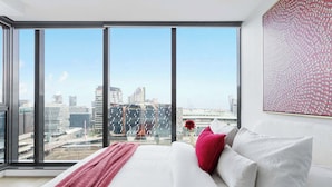 Une chambre d’hôtel moderne dotée d’une grande fenêtre donnant sur la ville, d’un lit bien fait avec du linge de lit blanc et un plaid rouge, ainsi que d’un élément décoratif mural.