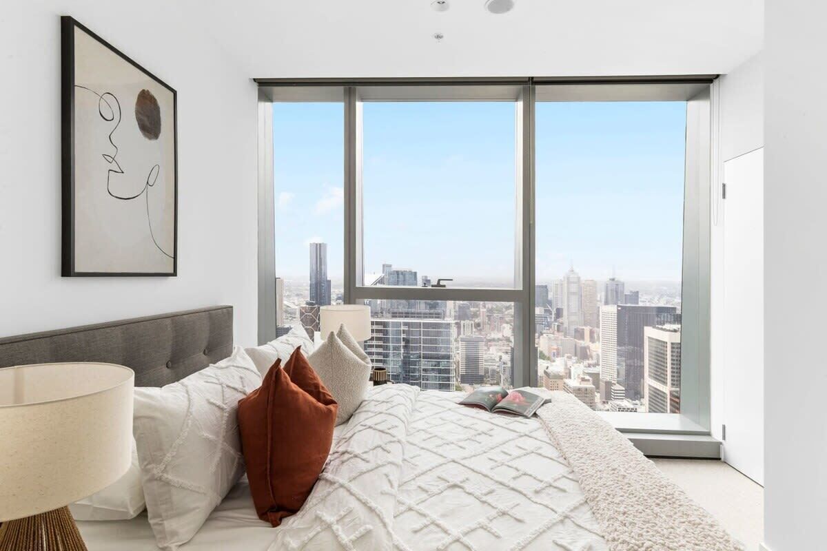 Ein modernes Schlafzimmer mit großem Fenster und Blick auf die Stadt, einem Bett mit weißer Tagesdecke und minimalistischer Einrichtung.