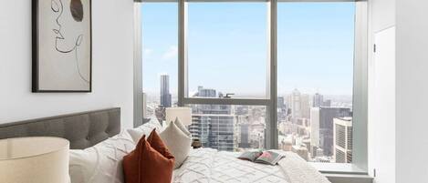 Ein modernes Schlafzimmer mit großem Fenster und Blick auf die Stadt, einem Bett mit weißer Tagesdecke und minimalistischer Einrichtung.