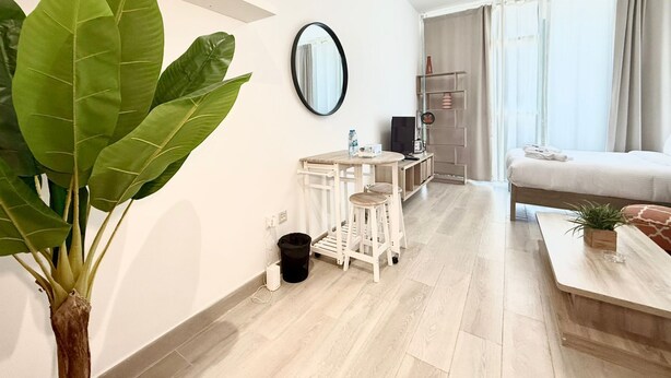 A modern, well-lit room with a large green plant, a round mirror, a small white desk, a bed with a white mattress, and a wooden coffee table.