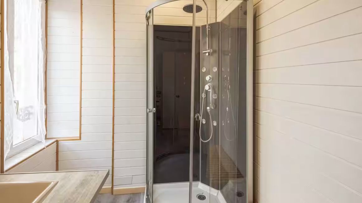 Una ducha con cerramiento de vidrio y puerta curva, suelo de madera y ventana a la izquierda.