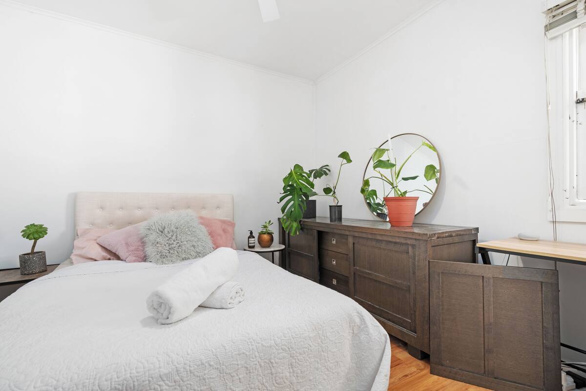 A bedroom with a bed, a dresser, a desk, and two potted plants.