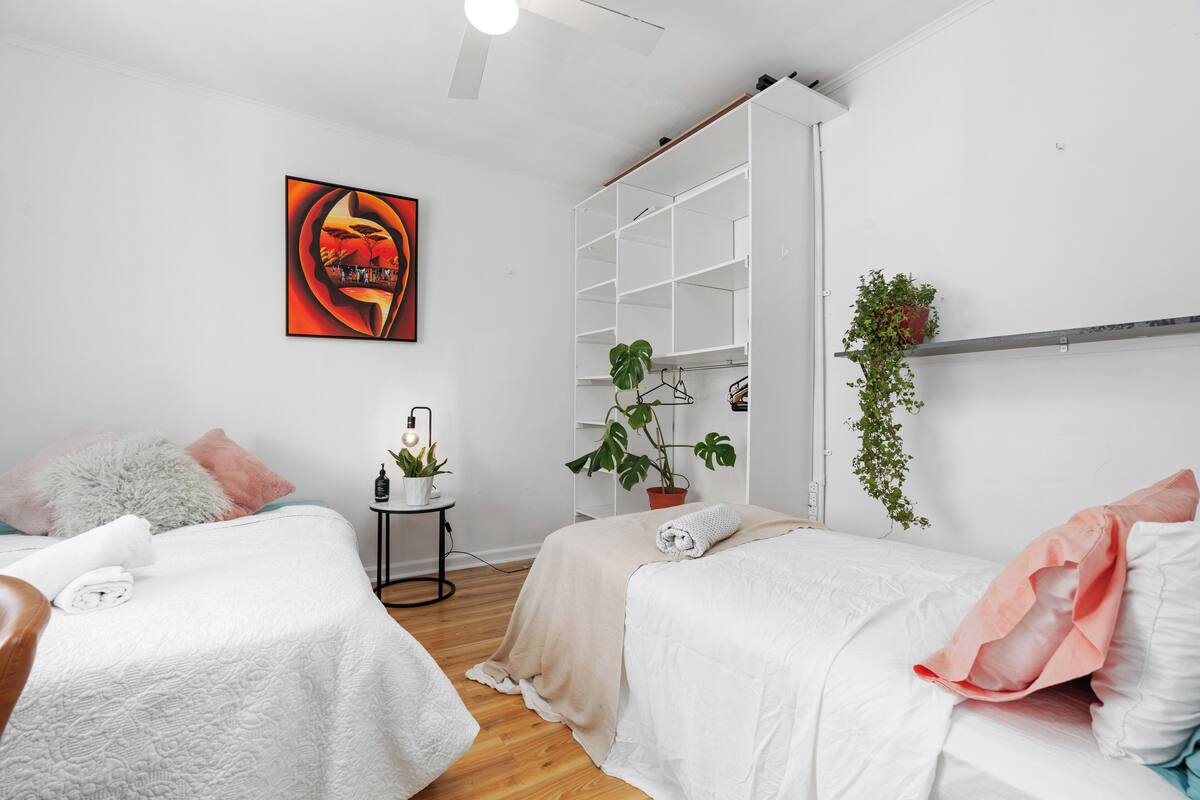 A modern bedroom with two beds, a large white wardrobe, a painting, and a small side table with a plant.
