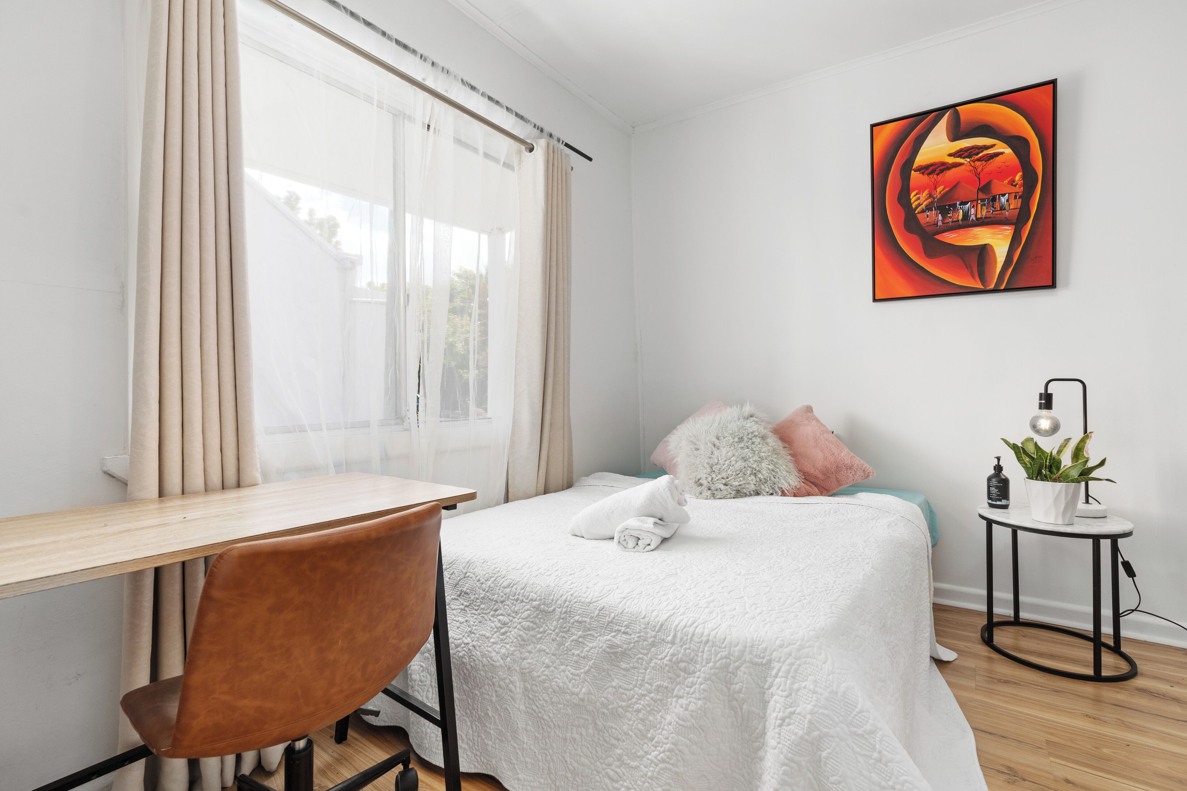 A bedroom with a bed, a desk, a chair, a bedside table with a lamp, and a painting on the wall.