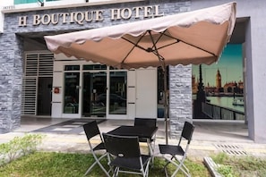 Een hotelentree met een bord waarop 'H BOUTIQUE HOTEL' staat, zitplaatsen buiten met een tafel en stoelen, en een uitzicht op een stadsbeeld met een brug.