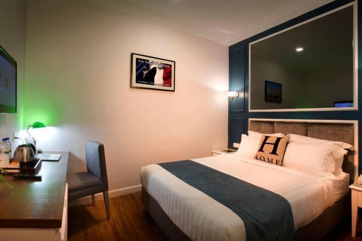 A hotel room with a bed, a desk with a chair, a framed picture on the wall, and a large mirror.