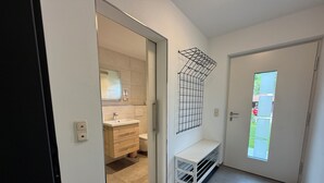 Une salle de bain moderne avec un lavabo, un miroir et des toilettes. On trouve un tapis noir devant la porte et un banc blanc avec des étagères.