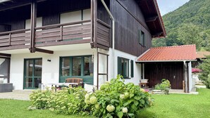 Une maison en bois à deux étages, dotée d’un balcon, d’un porche couvert et d’un jardin agrémenté d’hortensias.