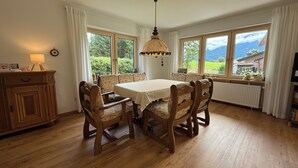 Une salle à manger avec une table et des chaises en bois, de grandes fenêtres avec des rideaux, un luminaire suspendu et un radiateur.
