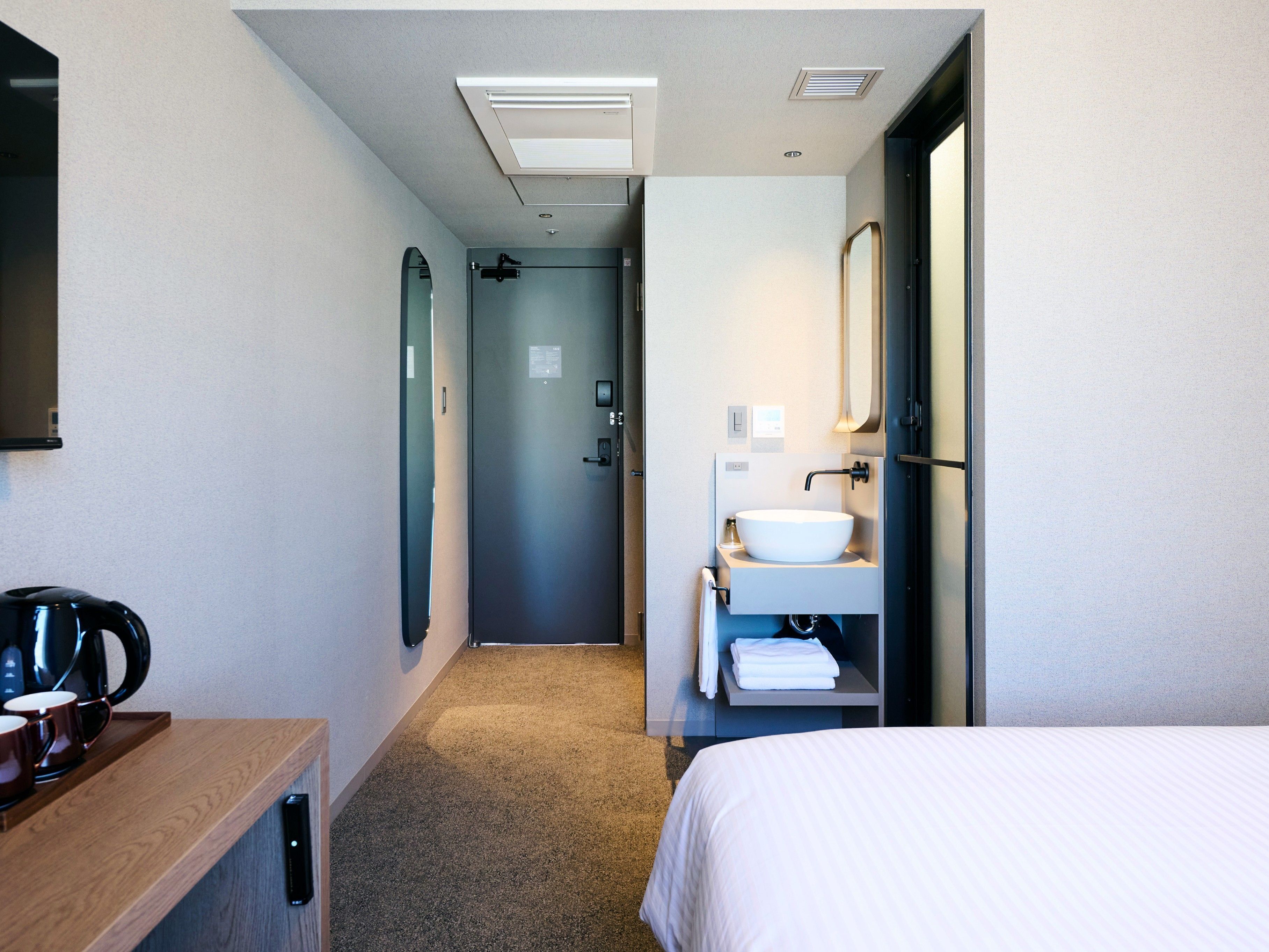 A hotel room with a bed, a bedside table with a coffee maker, a bathroom with a sink and mirror, and a door leading to the hallway.