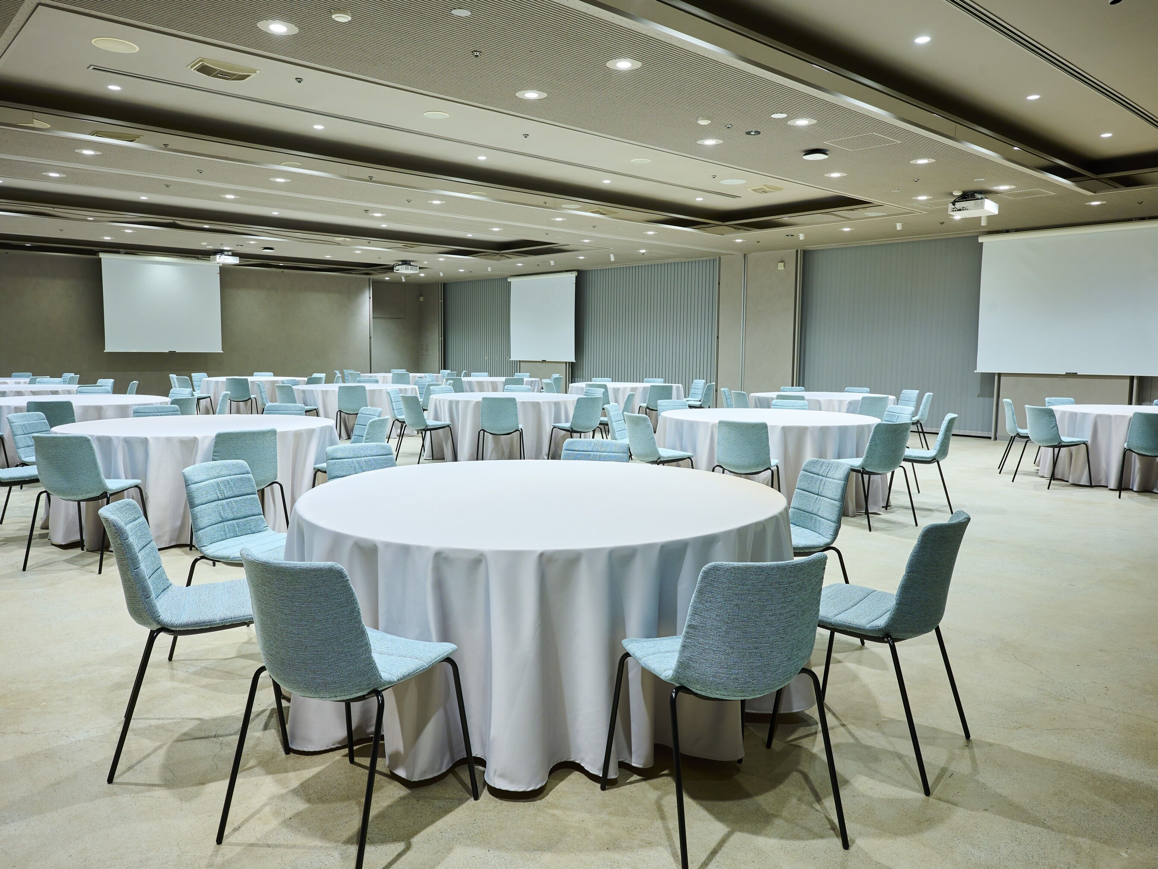 Un salón de conferencias con varias mesas redondas, cada una rodeada de sillas, y una pantalla grande al frente.