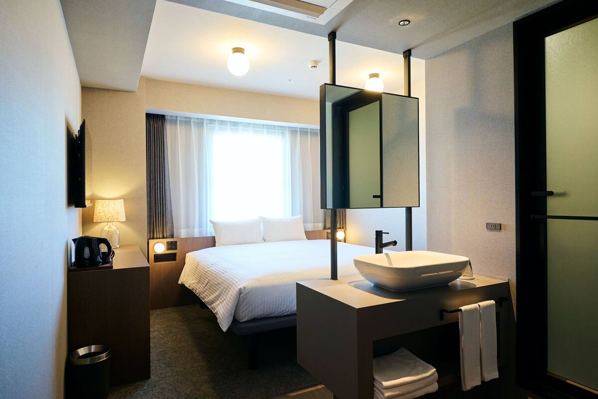 Ein Hotelzimmer mit Bett, Nachttisch, Lampe und einem großen Spiegel über einem modernen Waschbecken.