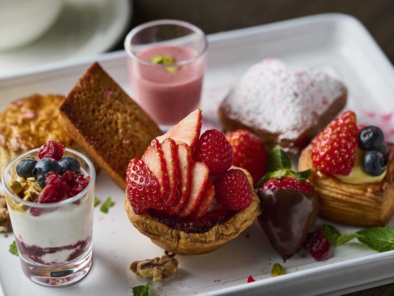 Uma bandeja de sobremesas com diversos itens, incluindo uma torta, uma fatia de bolo, um copo de bebida rosa e um pequeno pote de iogurte com frutas vermelhas.