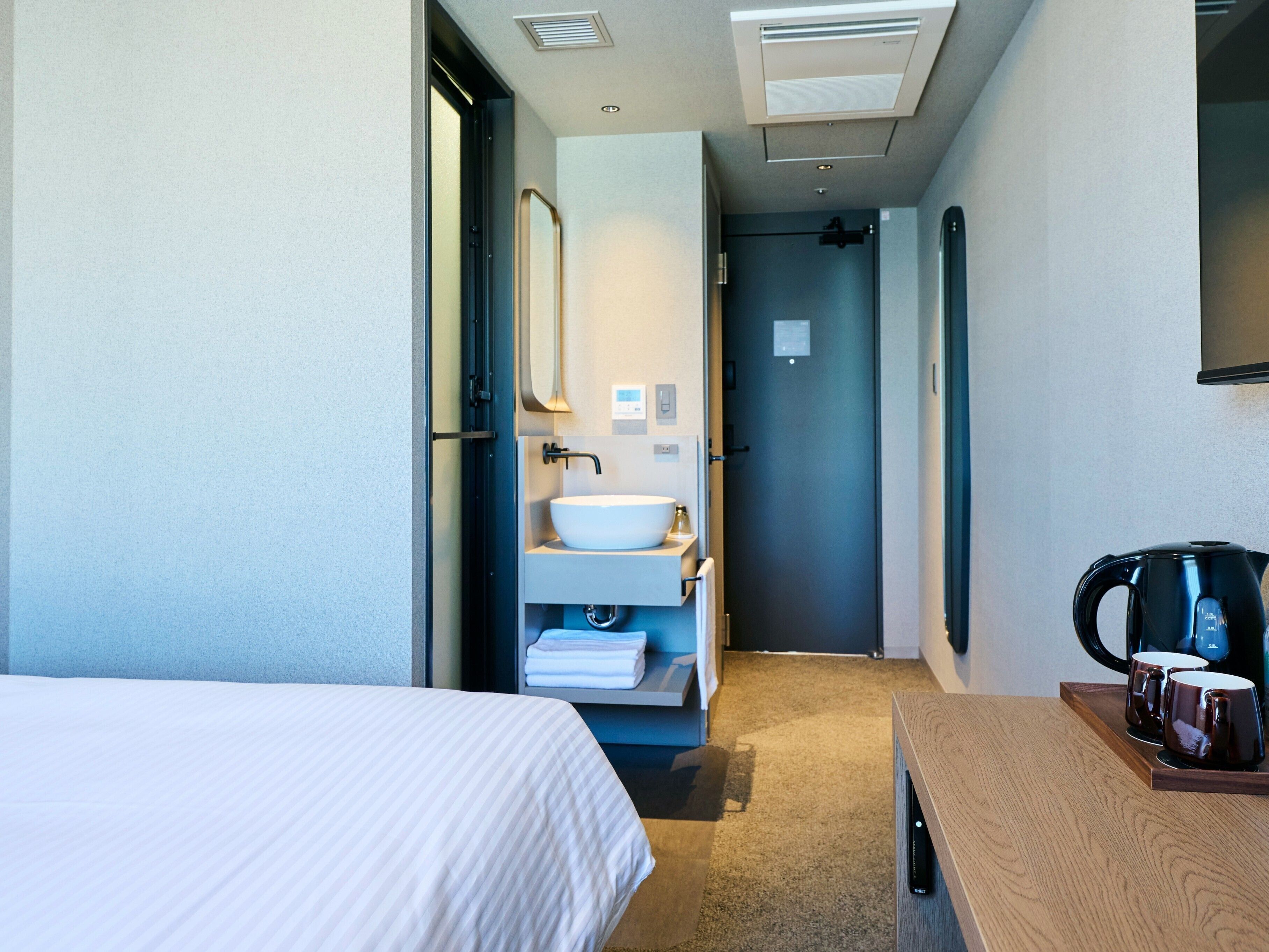 A hotel room with a bed, a wooden side table with a coffee maker and mugs, a bathroom with a sink and mirror, and a door leading to another room.