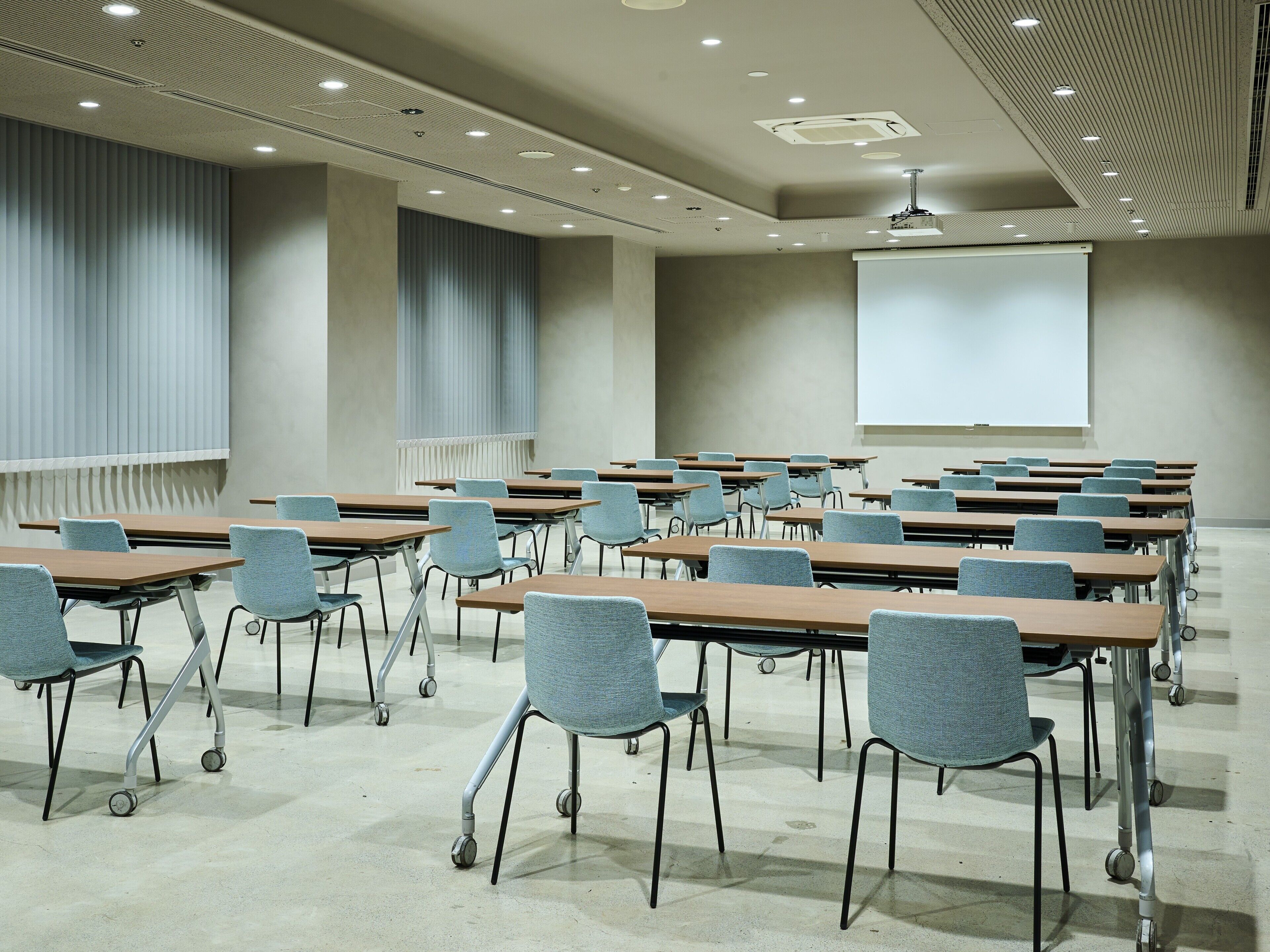 A conference room with rows of tables and chairs, a projector screen, and ceiling lights.
