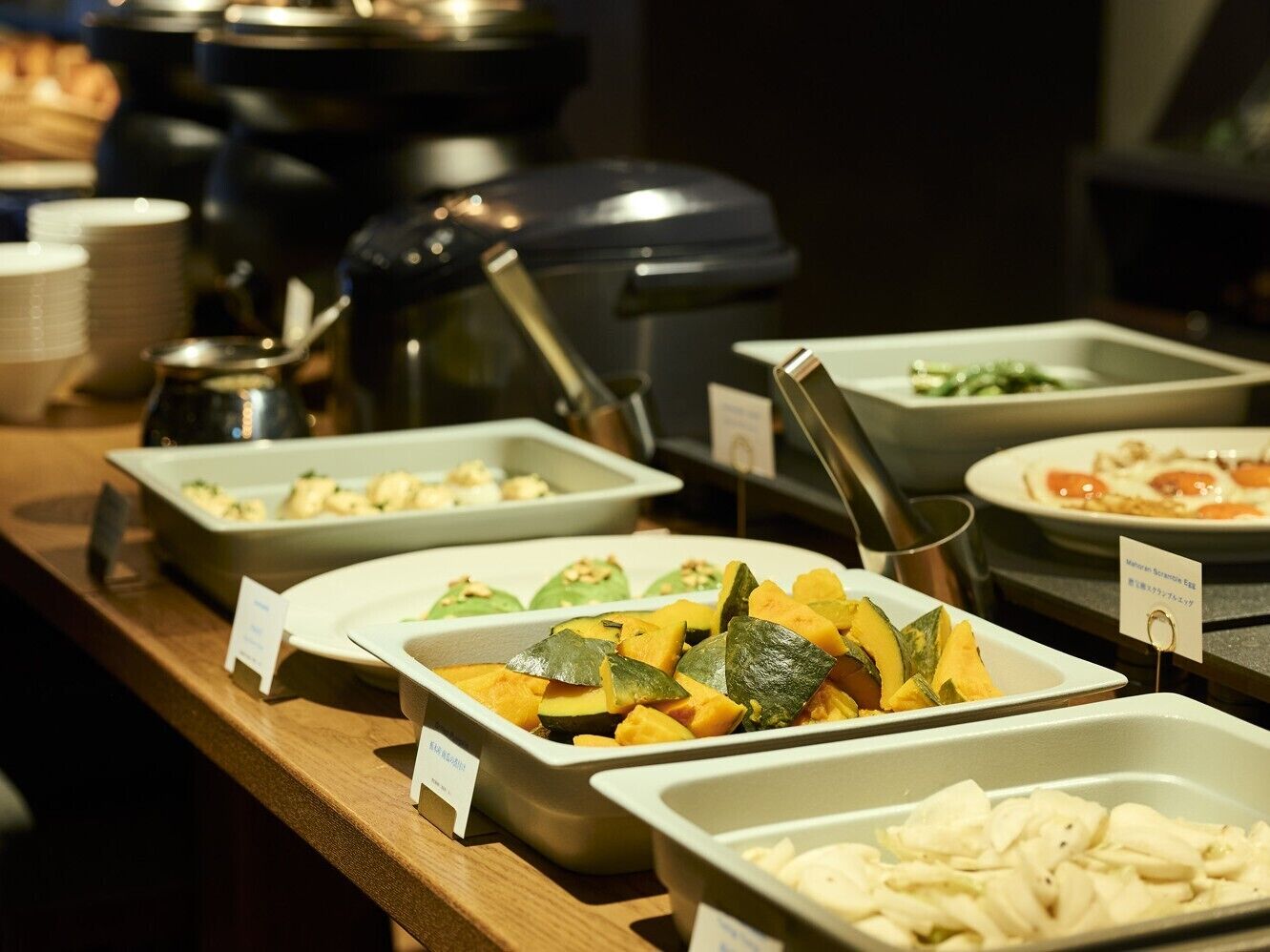 A buffet with various food options, including a dish with sliced vegetables and a container with a white creamy substance.