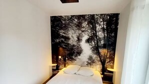 A bedroom with a bed, a wall-mounted lamp, and a large forest-themed painting. - Les suites du jardin 4 (Chaumont)