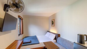 A compact room with a bed, a small table, a chair, a wall-mounted fan, a TV, and a kettle. - Andewi Cabin (Mengwi)