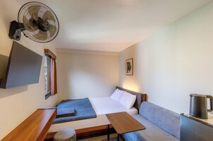 Una habitación pequeña con cama, una mesita, una silla, ventilador de pared, televisión y hervidor.