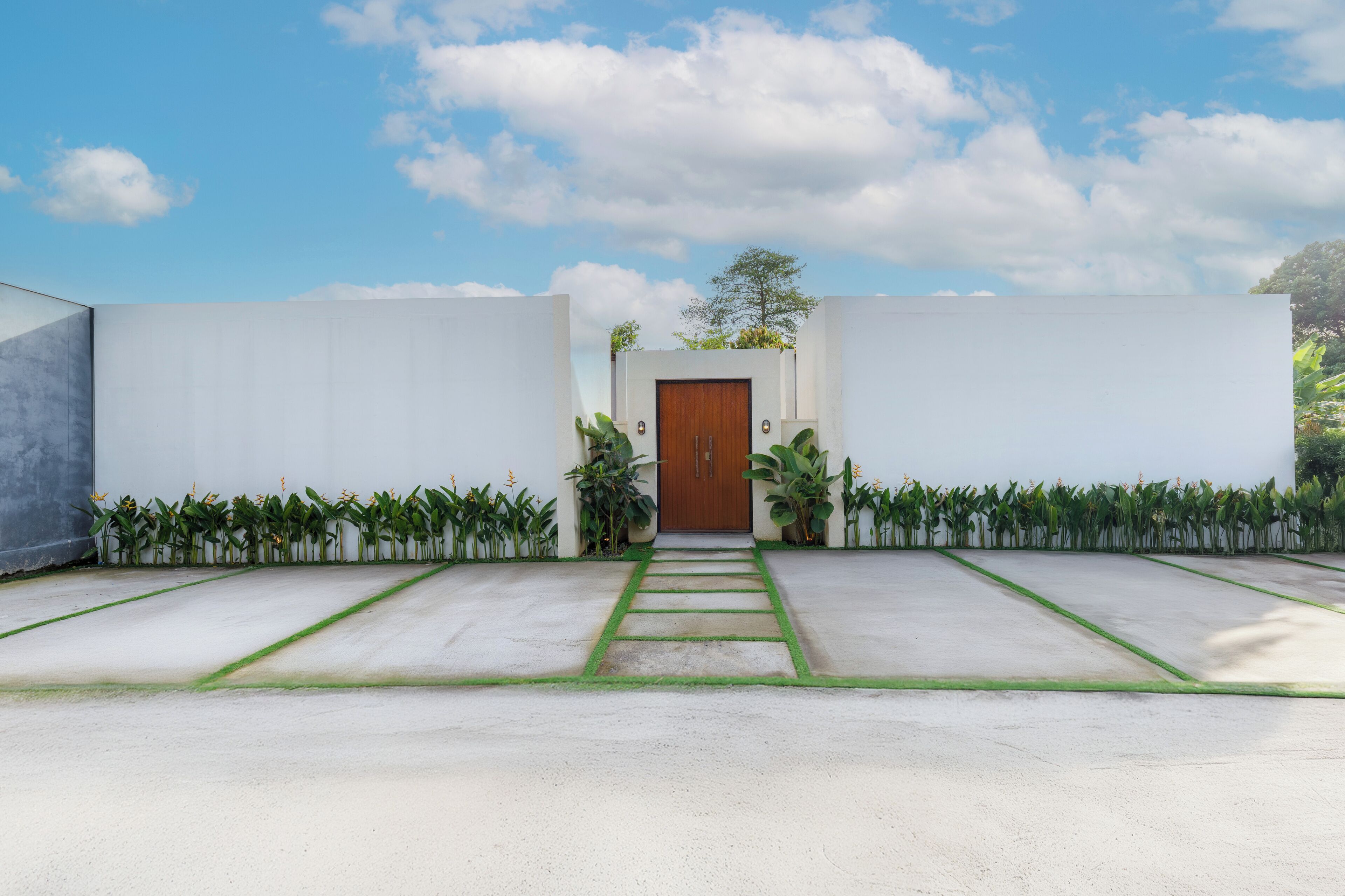 Un bâtiment moderne doté d’une porte centrale en bois, entouré d’un chemin en béton et bordé de plantes vertes.