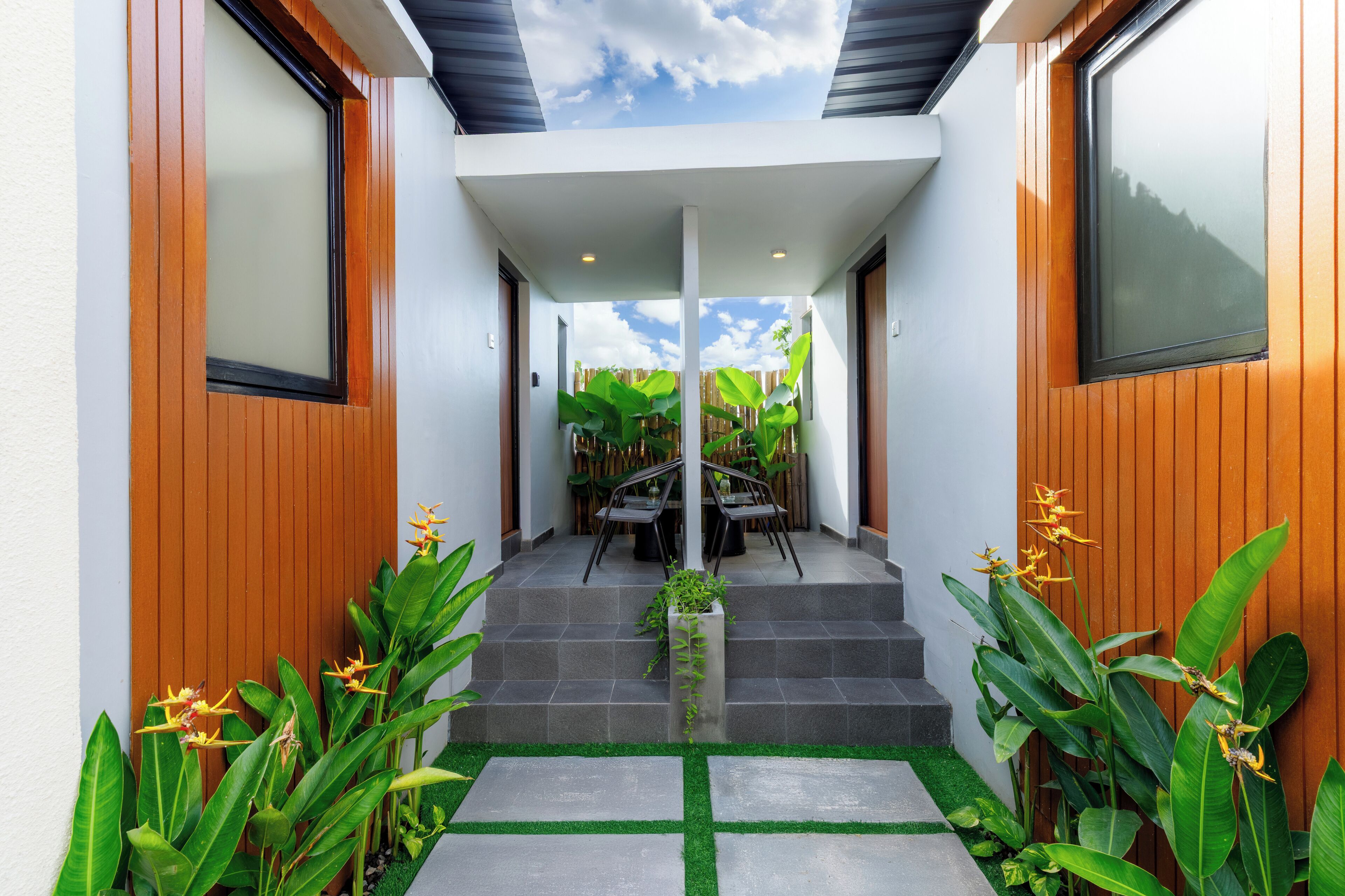 Una casa moderna con fachada de madera, una escalera que conduce a un porche cubierto y plantas tropicales.