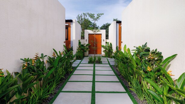 A modern pathway leading to a wooden door, flanked by lush greenery and white walls. - Andewi Cabin (Mengwi)