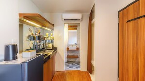 A narrow hallway with a kitchen area on the left, featuring a coffee machine, sink, and various kitchen utensils. A wooden door is on the right, and an air conditioning unit is mounted on the wall. - Andewi Cabin (Mengwi)