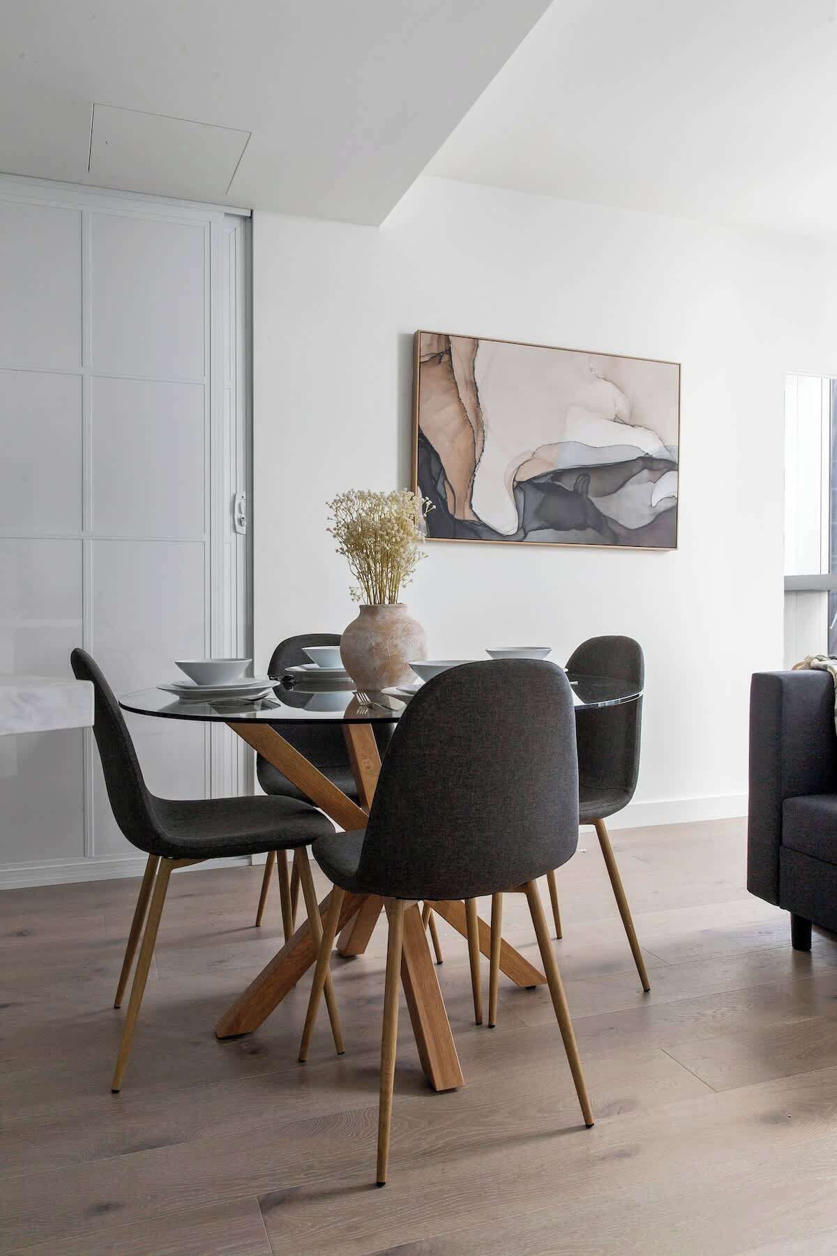 Un comedor moderno con una mesa de vidrio, sillas oscuras con patas de madera y un jarrón con flores secas.