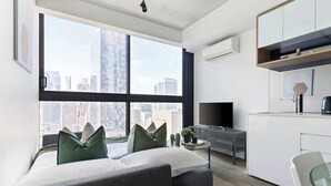 A modern apartment with a large window offering a city view, a kitchenette with a TV, and a seating area with cushions. - Pet-friendly Stylish 2BR Apt, Panoramic City Views (Melbourne)