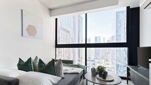 A modern living room with a large window, a grey sofa, and a small round table with a tray and potted plants. - Pet-friendly Stylish 2BR Apt, Panoramic City Views (Melbourne)