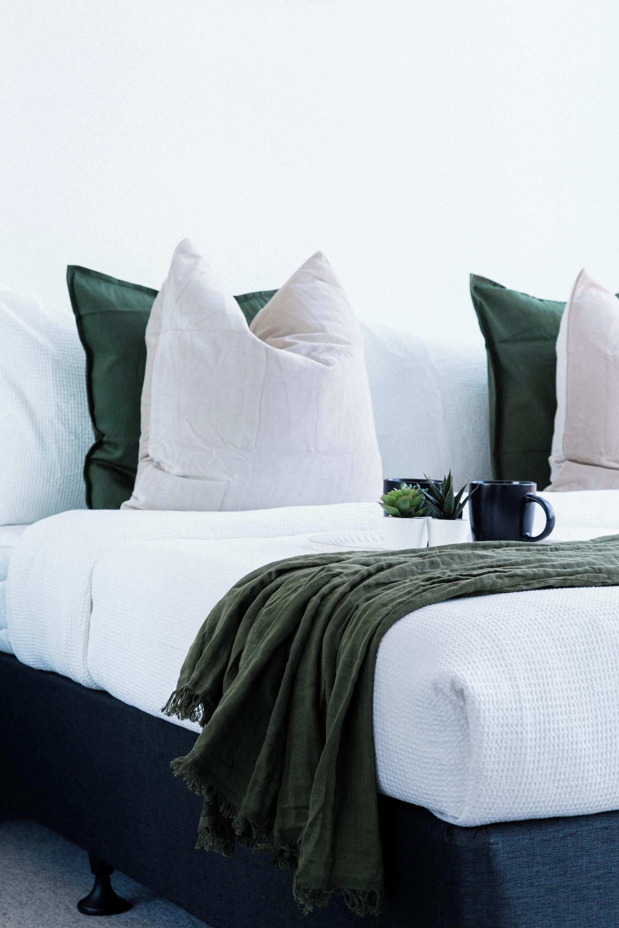 Una cama bien tendida con ropa de cama blanca y verde, un cabecero azul oscuro y una pequeña planta en maceta en la mesita de noche.