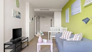 Una sala de estar con un televisor sobre un soporte negro, una mesa blanca con un florero, un sofá gris y una pared verde con cuadros.