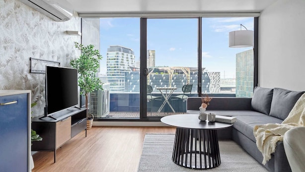 A modern living room with a large window, a flat-screen TV, a sofa, a round coffee table, and a small dining area with chairs. - Chic Apartment by Southern Cross, Free Parking (Melbourne)