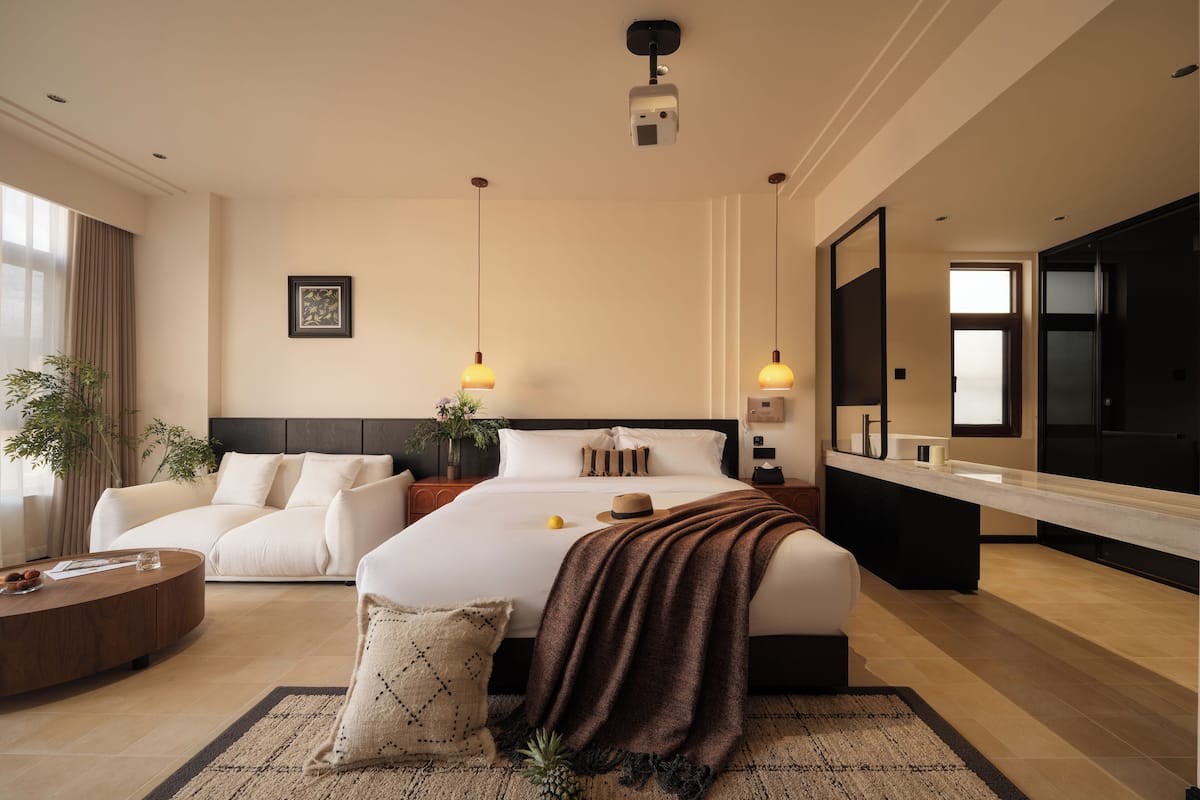 A modern hotel room with a large bed, a sitting area with a coffee table, a large mirror, and a view of the outside.