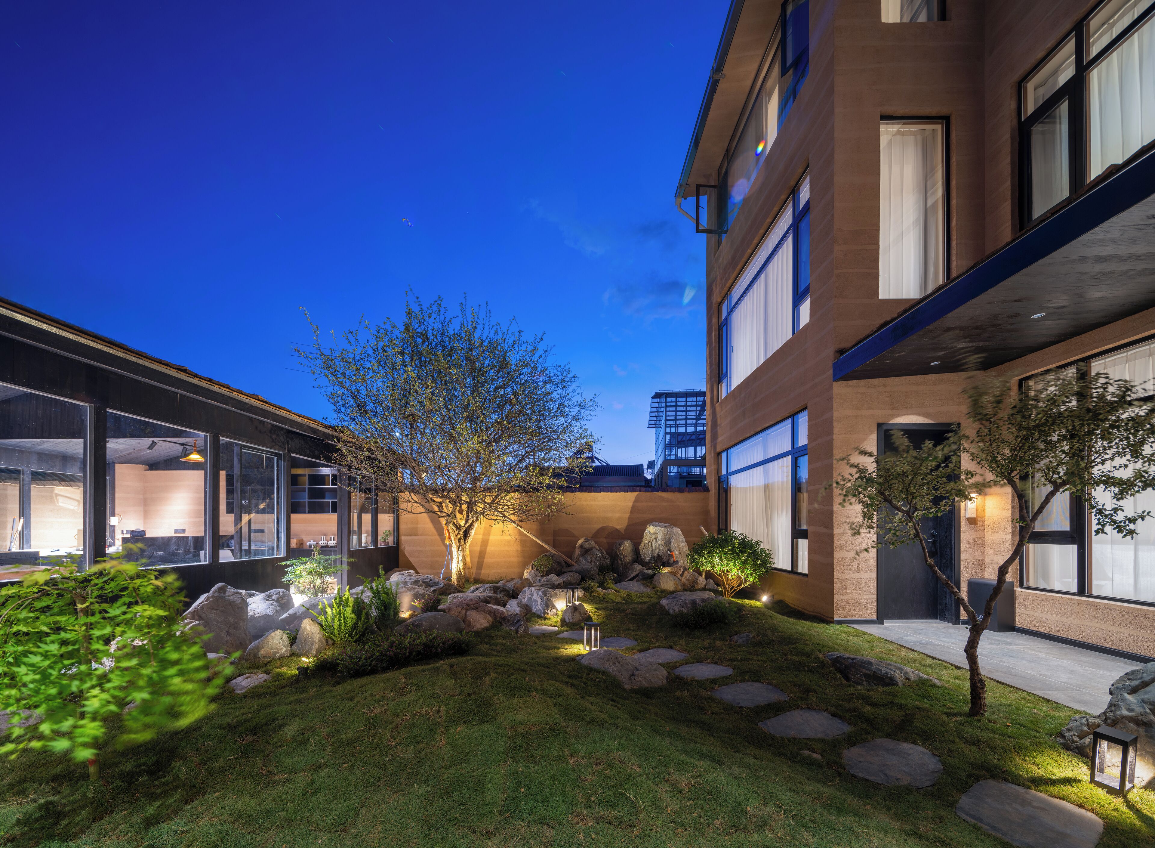 A modern building with large windows, a well-maintained garden with rocks and plants, and a clear blue sky at dusk.
