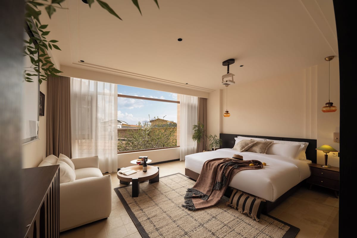 A modern hotel room with a large bed, a sofa, a small round table, and a view of the city through large windows.