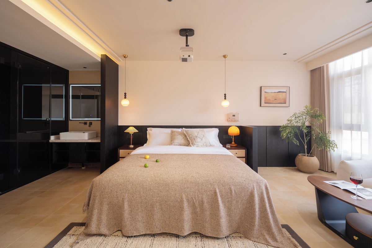 A modern hotel room with a large bed, bedside lamps, a bedside table, a potted plant, and a window with curtains.
