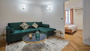 Un salon moderne avec un canapé vert, une table basse ronde et une petite table d’appoint. On aperçoit un lit avec une tête de lit rose au fond.