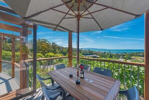 Een houten terras met een tafel en stoelen, een fles wijn en uitzicht op zee en groen.