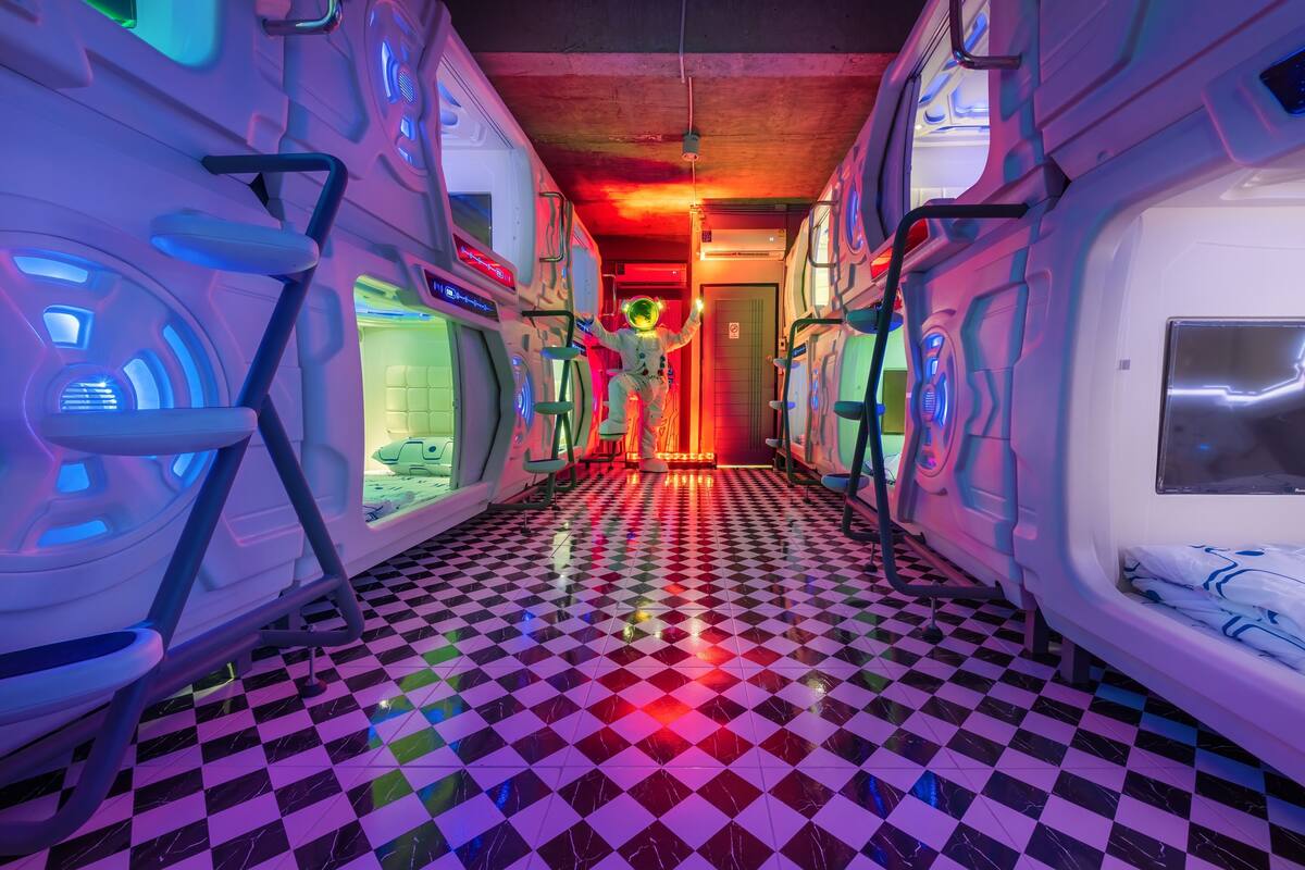 A futuristic hallway with checkered flooring and illuminated walls, leading to a room with a bed.