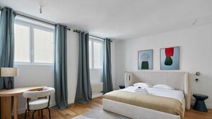 A modern bedroom with a bed, a desk, and a chair. There are two windows with curtains, a wall with two paintings, and a radiator. - Luxurious apartment Louvre Museum-Palais Royal (Paris)