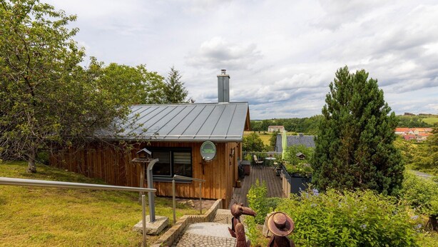 A rustic cabin with a metal roof, wooden walls, and a chimney. It features a stone staircase leading to a wooden deck with outdoor seating. - Holiday Home in Thale - Neinstedt (Thale - Neinstedt)