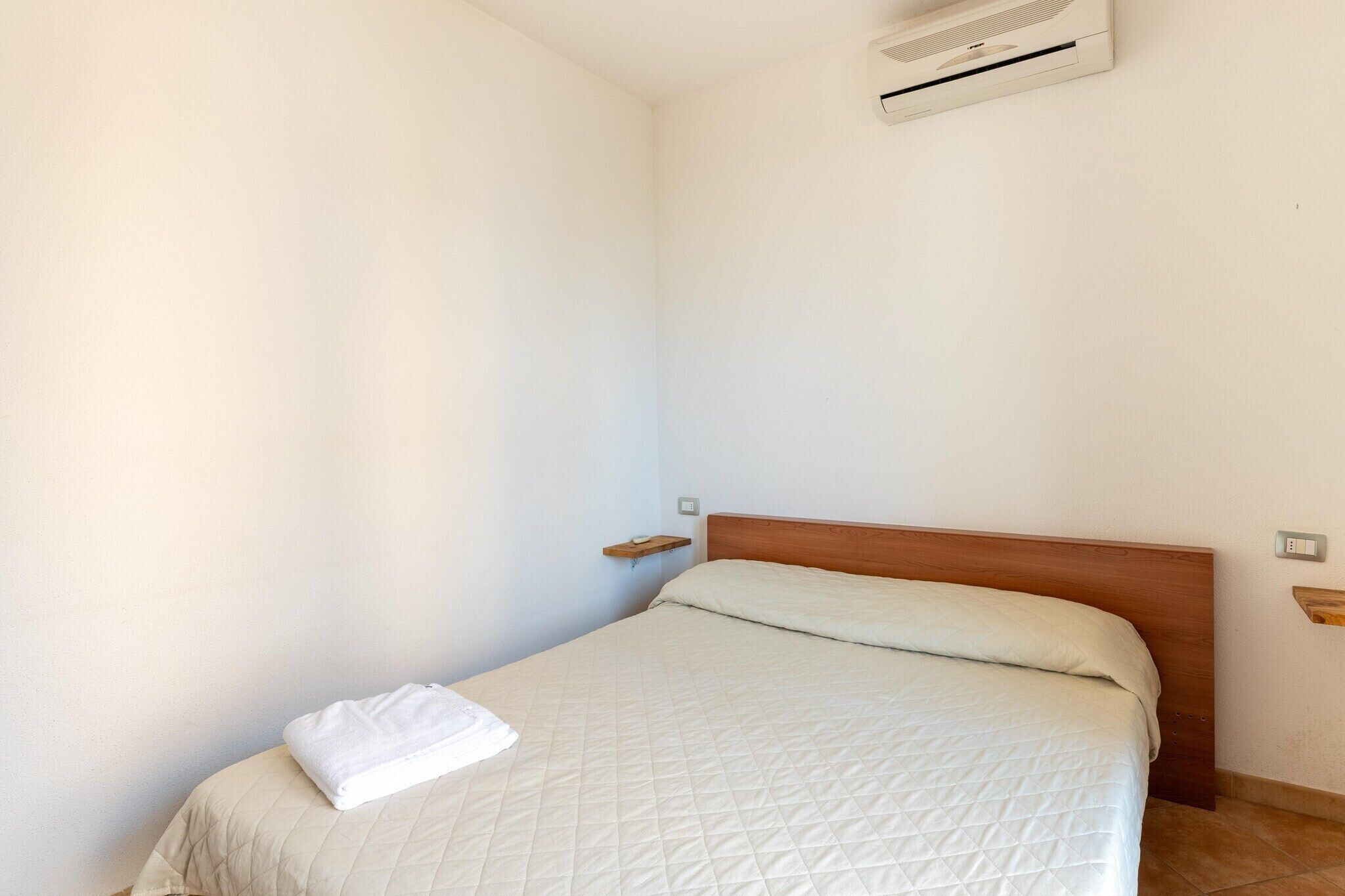A single bed with a wooden headboard, a white quilted bedspread, and a white towel on the side. There is a small wooden shelf on the wall to the left of the bed and a wall-mounted air conditioning unit above it.
