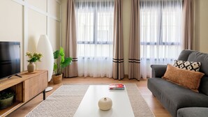 Een moderne woonkamer met een flatscreen televisie, een bank, een salontafel en een groot raam met dunne gordijnen.